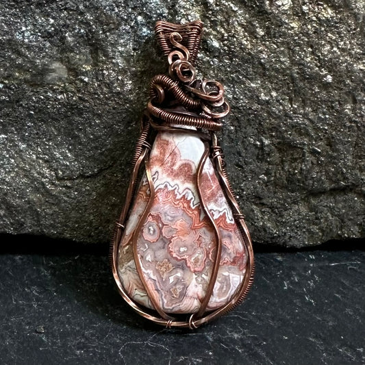 A teardrop-shaped copper wire-wrapped pendant crazy lace agate stone, set against a dark textured rock background.