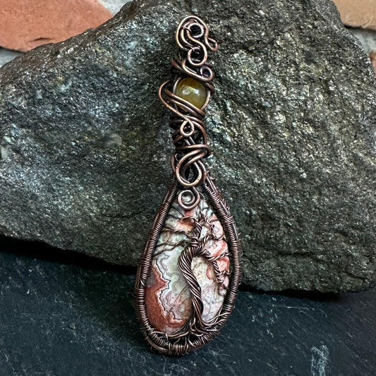 A handmade copper wire wrapped teardrop pendant featuring a tree-of-life design on a piece of crazy lace agate, wrapped in copper wire, with a small olive green bead at the top, displayed against a dark textured rock.
