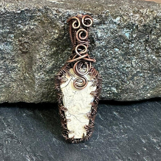 A handmade copper wire pendant with a white coffin shaped Maligano jasper stone, resting against a textured gray rock on a dark slate surface