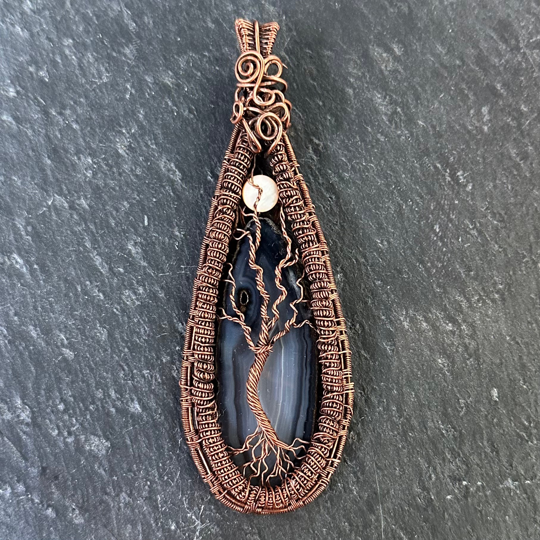 A teardrop-shaped pendant featuring a dark black-gray agate stone with a copper wire tree design, set against a dark gray slate background.