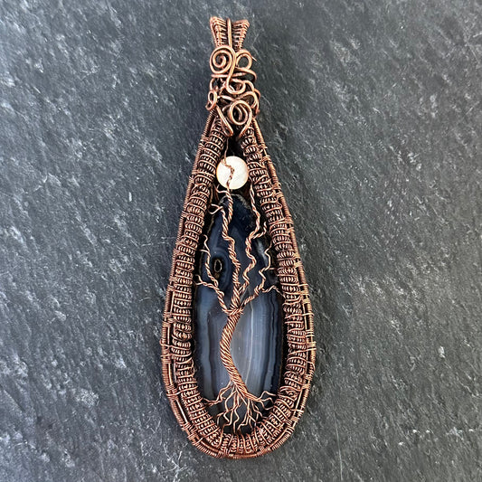 A teardrop-shaped pendant featuring a dark black-gray agate stone with a copper wire tree design, set against a dark gray slate background.