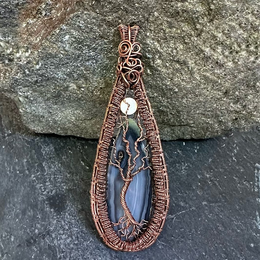 A teardrop-shaped pendant featuring a black agate stone with a copper wire tree of life design, resting against a textured gray rock.