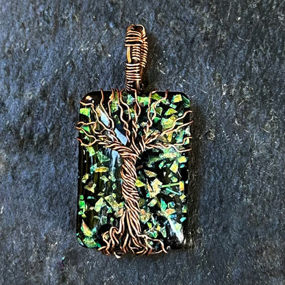 A rectangular pendant featuring a tree of life design wrapped in copper wire over a black and green iridescent mosaic background, shown on a dark textured stone surface.