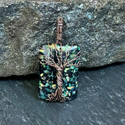 A handmade pendant featuring a square iridescent green and blue opal resin piece wrapped with a copper tree design, set against a dark textured stone background.