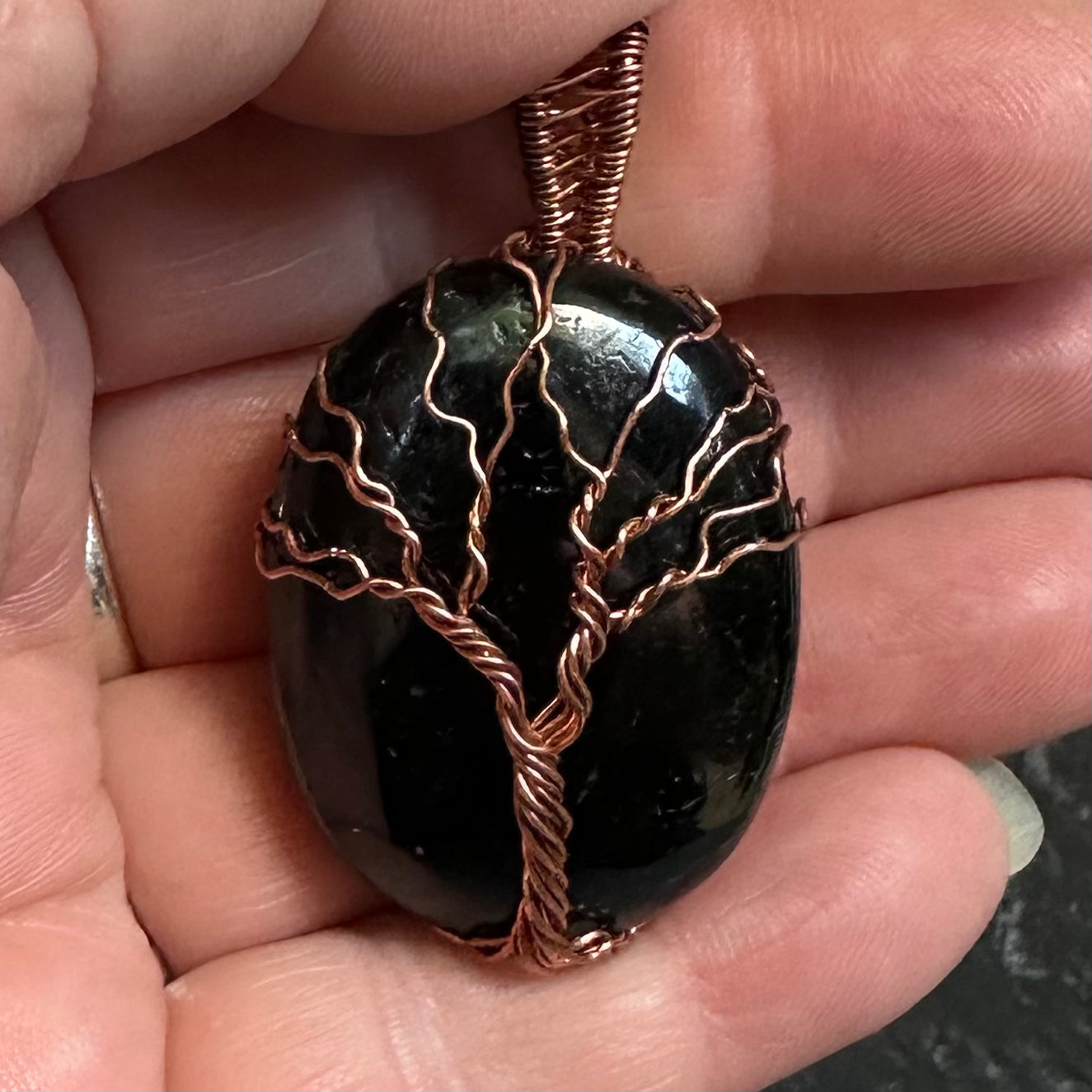 A close-up of a hand holding a polished oval black tourmaline pendant wrapped with a copper tree design.