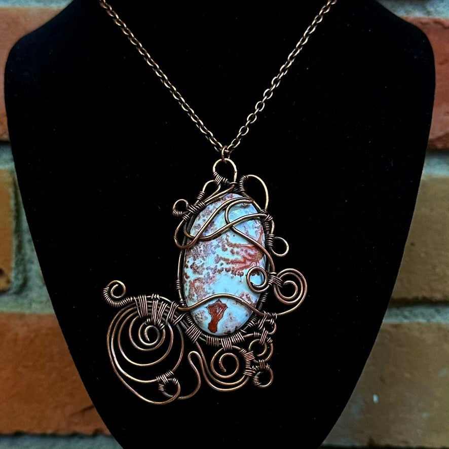 A handmade wire-wrapped necklace featuring crazy lace agate stone pendant, displayed on a black velvet jewelry bust against a blurred brick wall background.