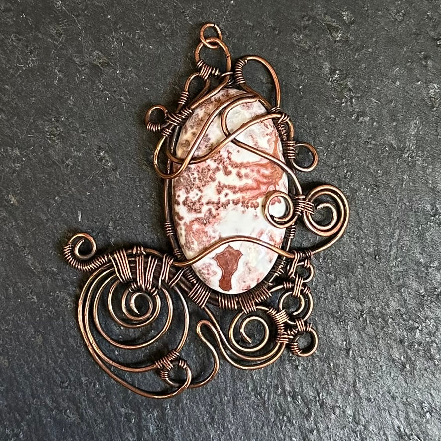 A handmade oval pendant featuring a an oval crazy lace agte cabochon wrapped in intricate copper wire with decorative swirls, displayed on a dark textured slate background.