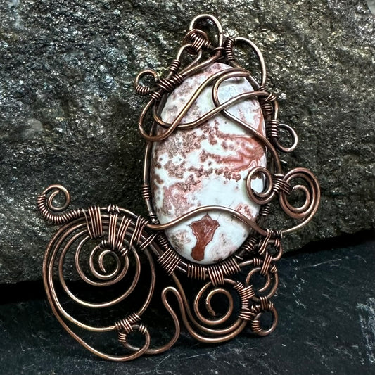 A handmade copper wire pendant featuring a crazy lace agate stone cabochon, set against a dark textured rock background.