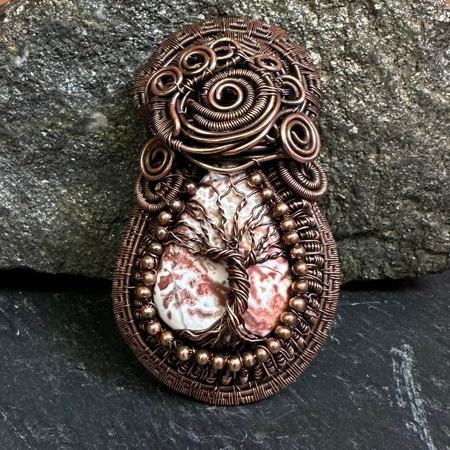 A copper wire-wrapped pendant featuring a central tree of life design over a crazy lace agate stone, displayed against a dark textured slate and rock background.