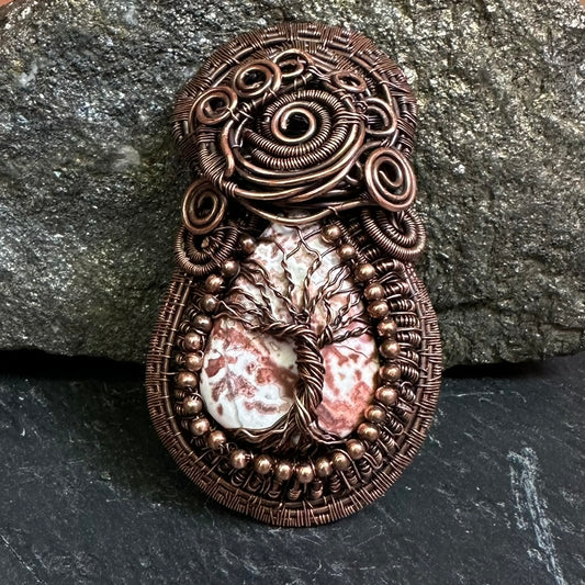 A copper wire-wrapped pendant featuring a central tree of life design over a crazy lace agate stone, displayed against a dark textured slate and rock background.