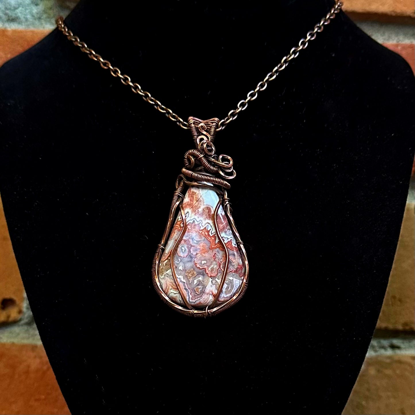 A copper wire wrapped pendant featuring a crazy lace agate stone displayed on a black jewelry bust.