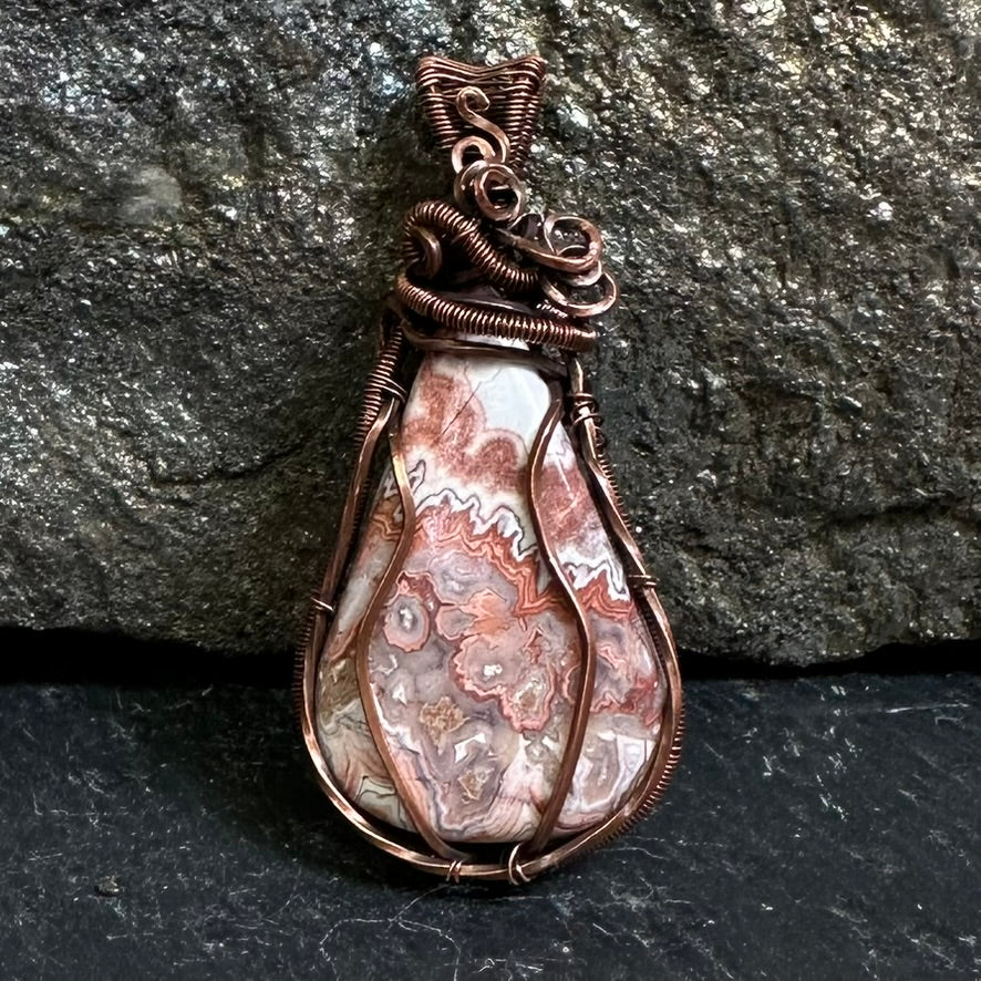 A teardrop-shaped copper wire-wrapped pendant crazy lace agate stone, set against a dark textured rock background.