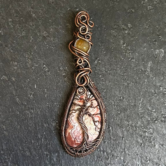A teardrop-shaped copper wire-wrapped pendant featuring a Tree of Life design set in crazy lace again stone, with a small round yellow-orange bead accent at the top, displayed on a dark textured slate surface.