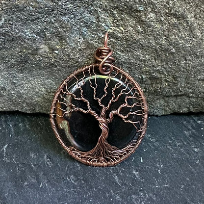 A circular tree of life pendant featuring a sardonyx center and a copper wire wrap design, displayed against a textured gray stone background.