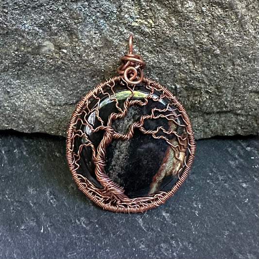 A handmade oval pendant featuring a copper wire tree of life design over a sardonyx stone, set against a dark gray slate and textured rock background.