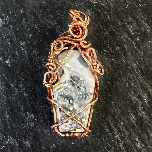 A hand-wrapped copper wire pendant featuring a gray Maligano jasper stone with natural inclusions, set against a dark textured slate background.