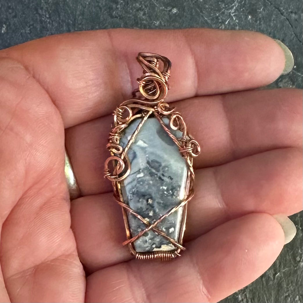 A copper wire wrapped coffin shaped grey Maligano jasper stone wrapped in copper wire held in a person's hand.