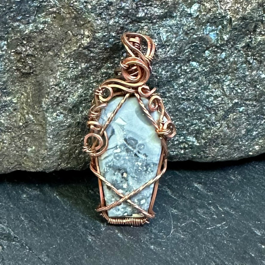 A coffin-shaped grey Maligano jasper pendant with a copper wire wrap, resting on a dark textured slate surface with a gray rock in the background.