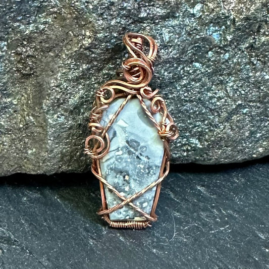 A coffin-shaped grey Maligano jasper pendant with a copper wire wrap, resting on a dark textured slate surface with a gray rock in the background.