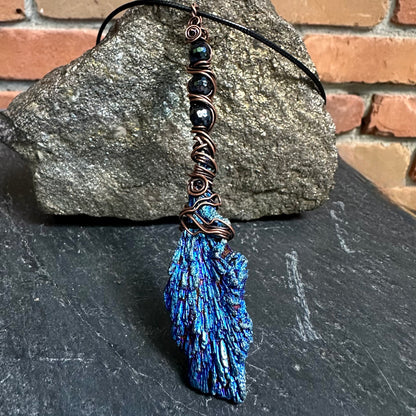A handmade pendant necklace featuring a textured blue and black iridescent kyanite stone wrapped in copper wire, suspended from dark beads on a black cord, displayed against a gray rock and brick background.