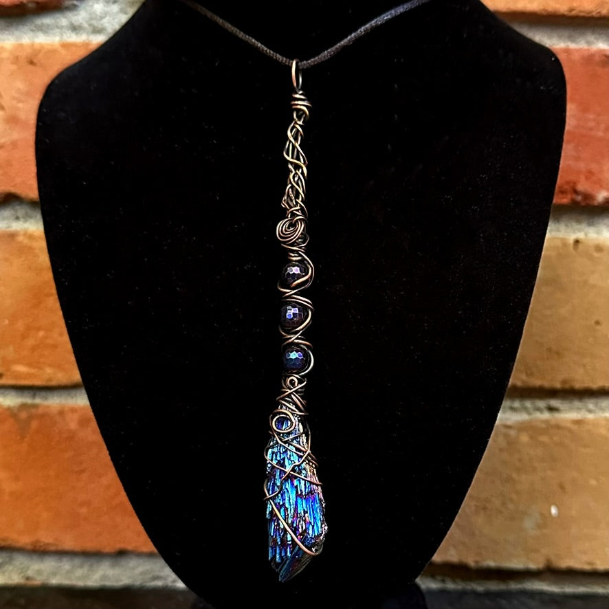 A handmade copper wire-wrapped pendant necklace featuring a blue iridescent kyanite stone and dark iridescent beads, displayed on a black velvet jewelry bust against a brick wall background.