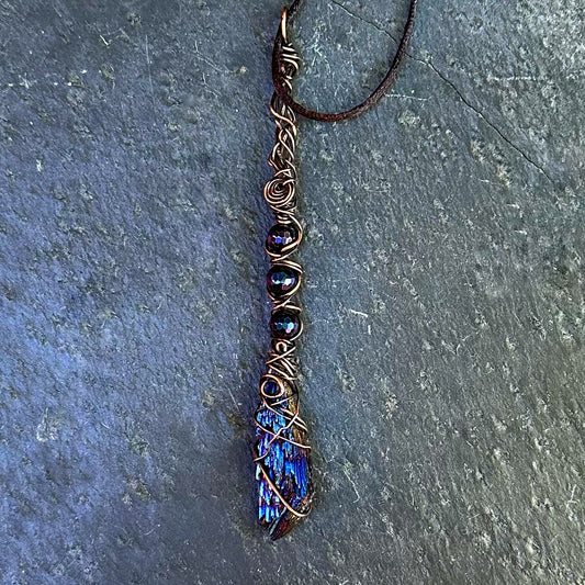 A handmade wire-wrapped pendant necklace featuring a blue iridescent kyanite stone and dark beads, wrapped in copper-toned wire and suspended from a dark cord, displayed on a textured slate surface.