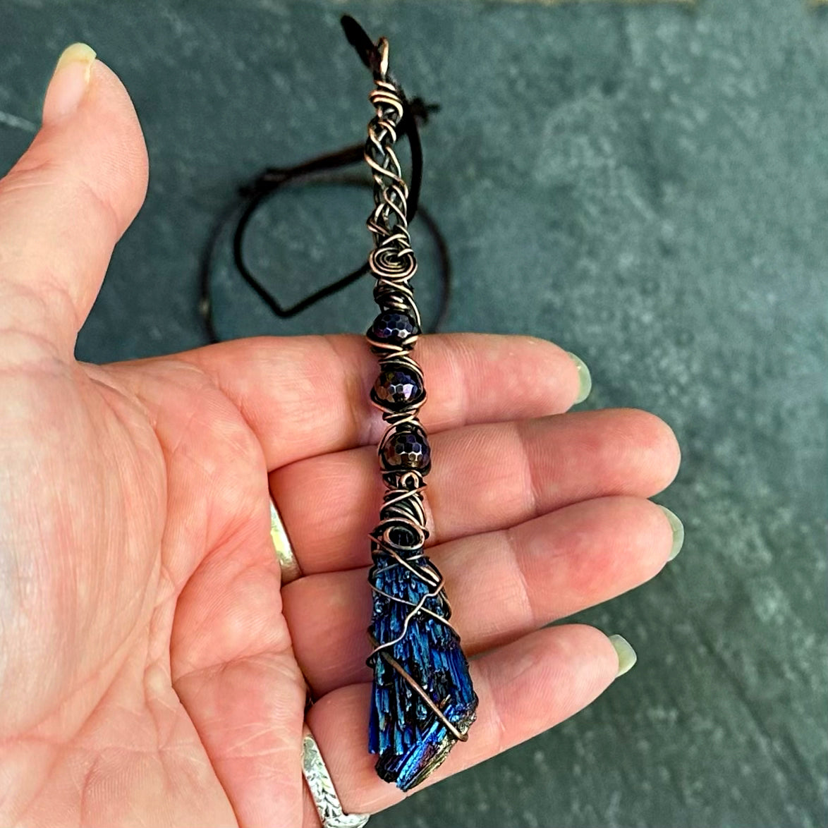 A hand holding a necklace featuring a large blue iridescent kyanite stone wrapped in copper wire, with dark faceted beads and a braided metal top section, set against a slate background.