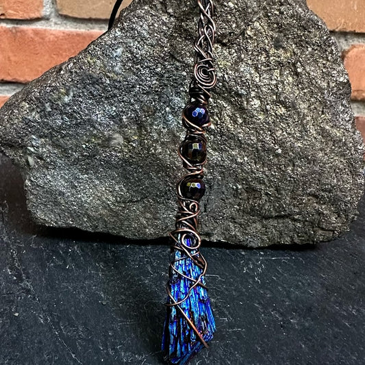 A handmade wire-wrapped necklace featuring dark faceted beads and a blue iridescent kyanite stone wrapped in copper wire, displayed against a dark gray rock with a brick wall background.