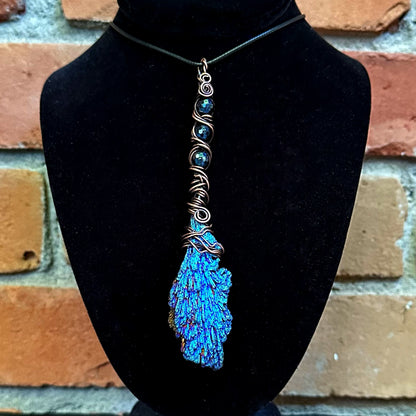 A handmade copper wire-wrapped pendant necklace featuring a vibrant blue iridescent kyanite stone, displayed on a black velvet jewelry bust against a rustic brick wall background.