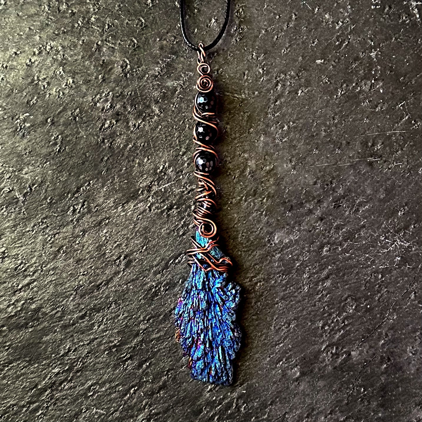 A handmade pendant necklace featuring a textured blue and turquoise kyanite stone wrapped in copper wire, accented with black beads and a black cord, displayed on a dark textured stone surface.