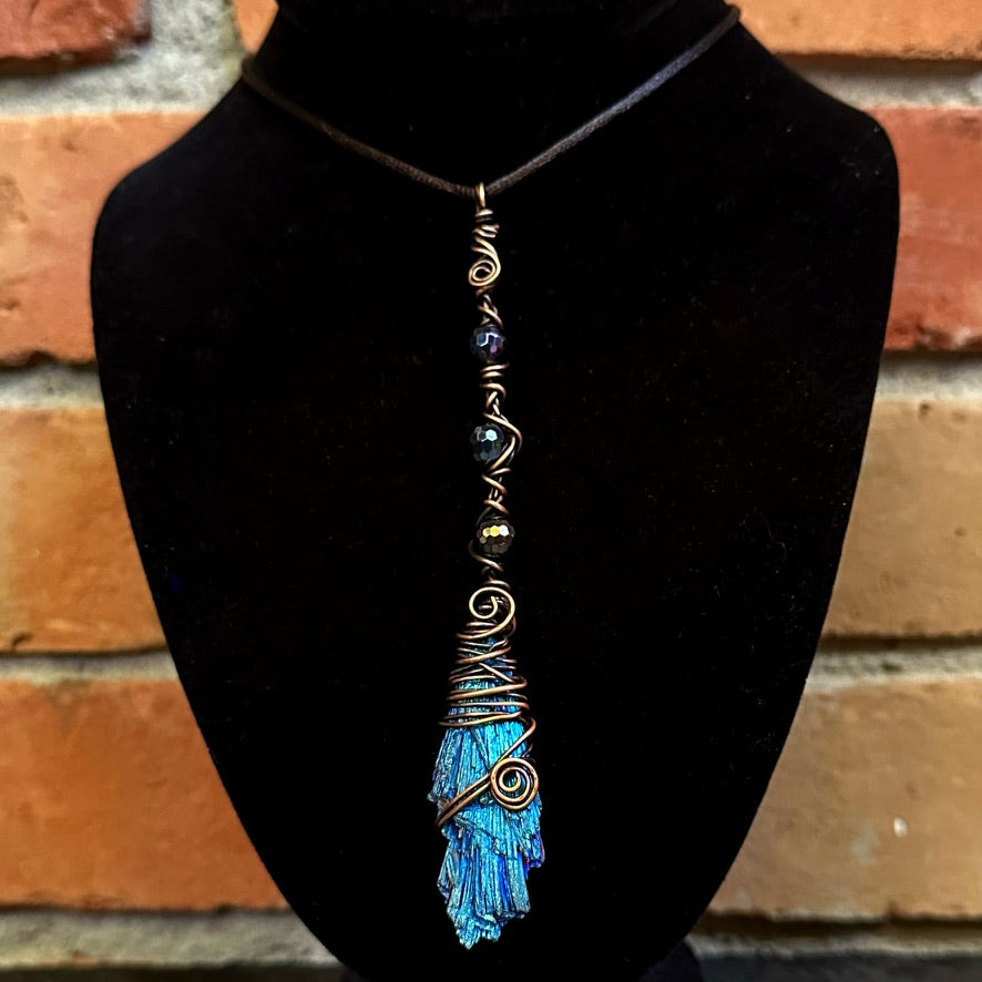 A handmade pendant necklace featuring a blue iridescent kyanite stone wrapped in copper wire, displayed on a black velvet jewelry bust against a brick wall background.