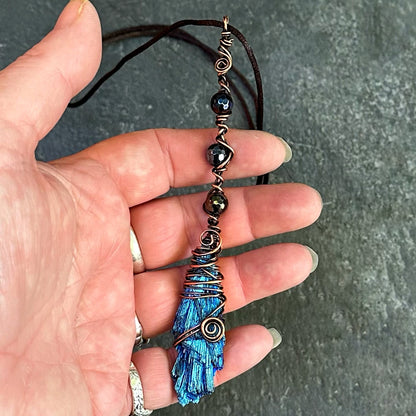 A handmade wire-wrapped pendant necklace held in a hand. The necklace features iridescent beads, and a large blue iridescent kyanite stone wrapped in copper-toned wire.