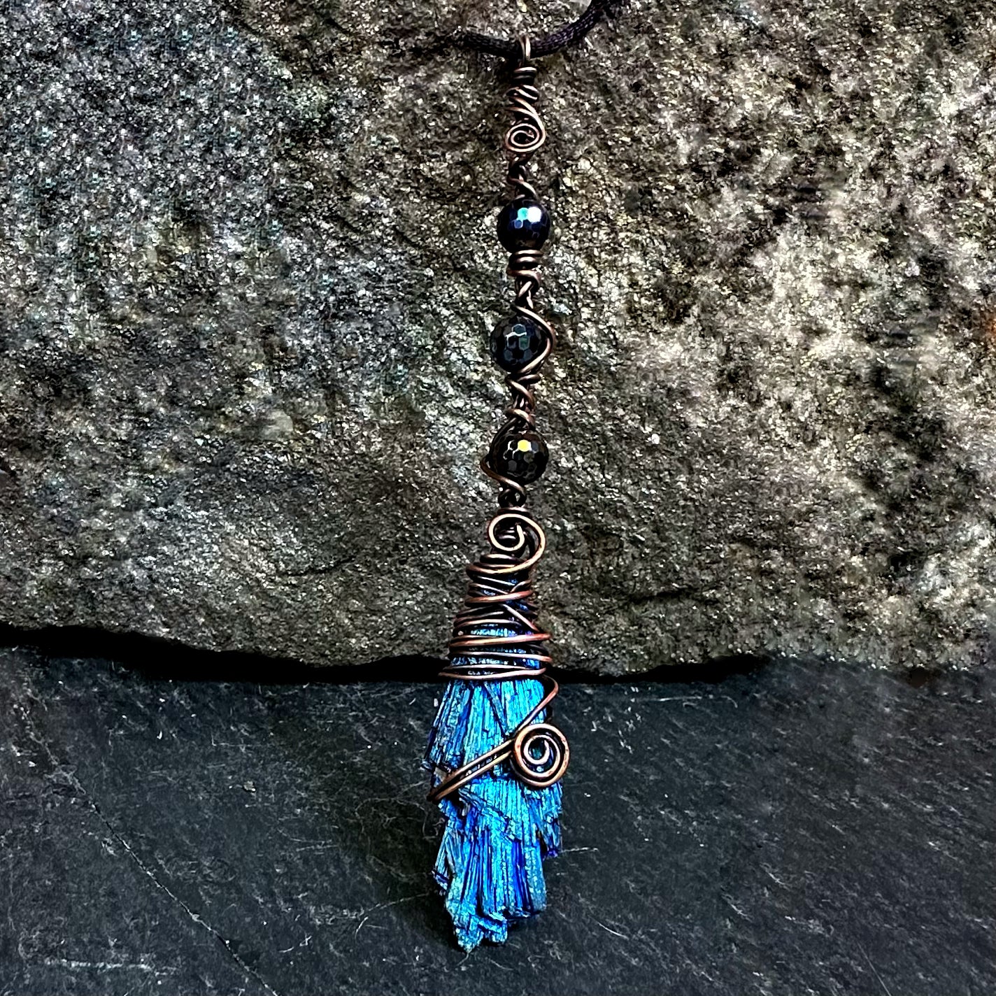A handmade wire-wrapped kyanite stone necklace featuring black beads, copper wire spirals, displayed against a dark textured stone background.