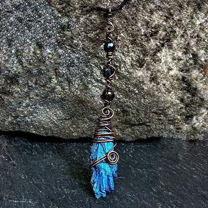 A handmade wire-wrapped kyanite stone necklace featuring black beads, copper wire spirals, displayed against a dark textured stone background.