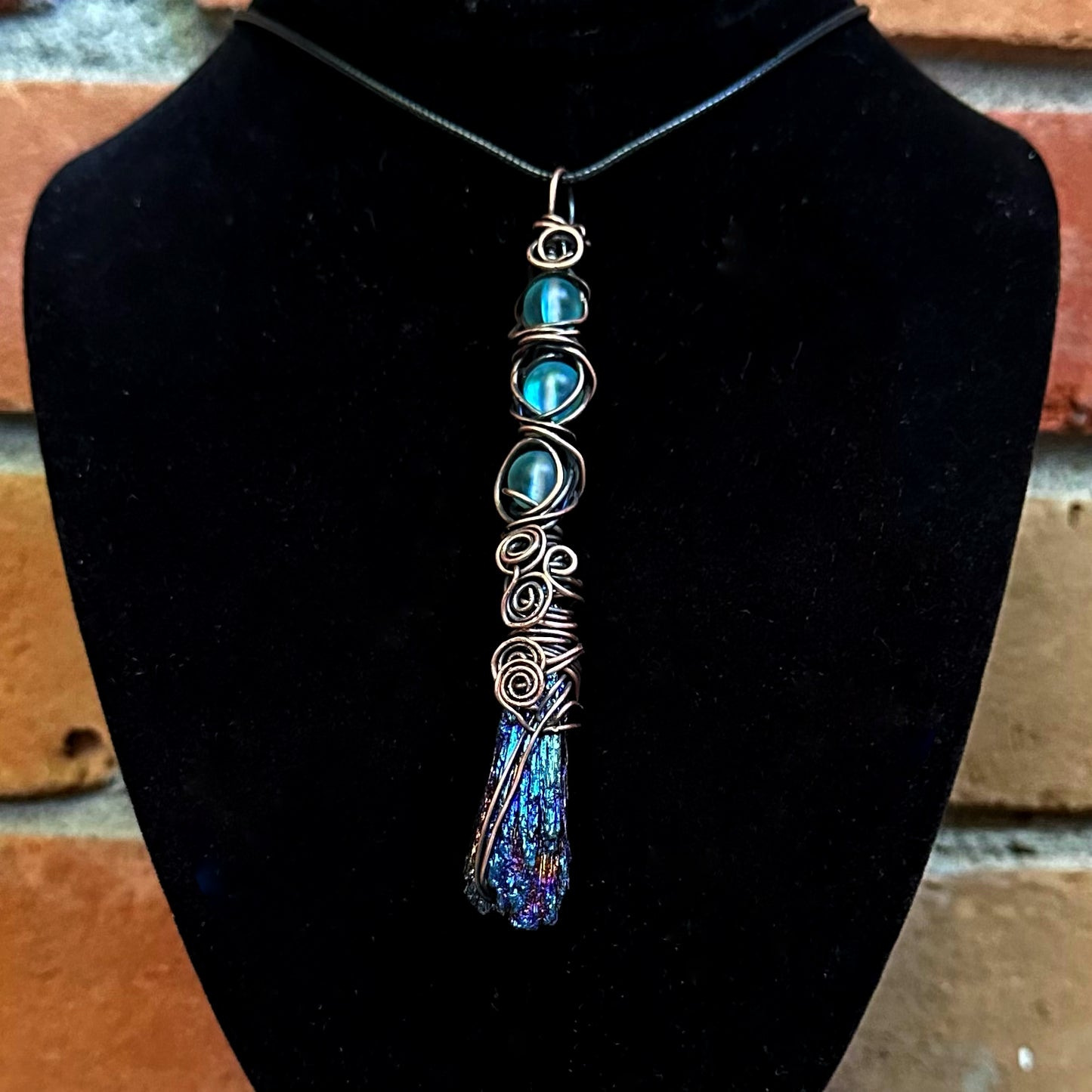 A handmade copper wire-wrapped necklace featuring a titanium aura kyanite stone, displayed on a black velvet jewelry bust against a brick wall background.