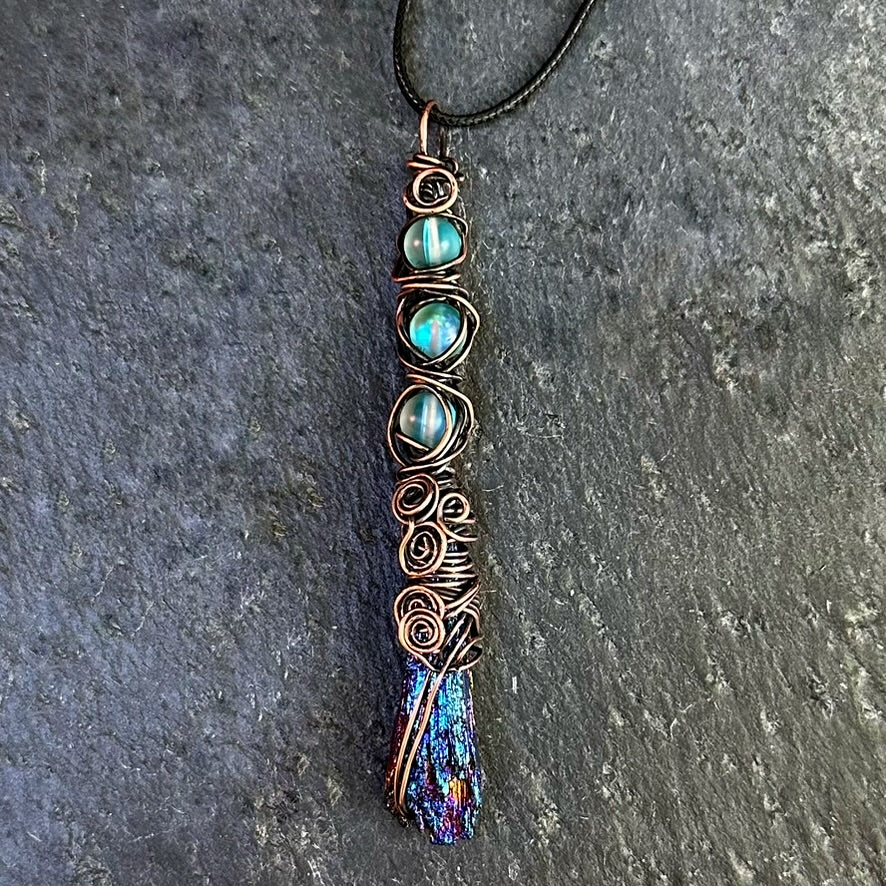 A handmade copper wire-wrapped pendant necklace featuring iridescent blue beads and an iridescent kyanite stone, shown on a dark gray textured stone background.