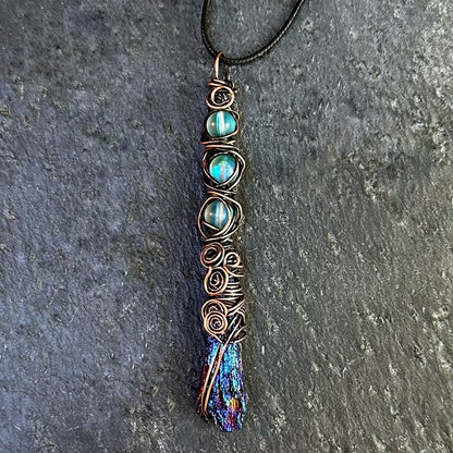 A handmade copper wire-wrapped pendant necklace featuring iridescent blue beads and an iridescent kyanite stone, shown on a dark gray textured stone background.