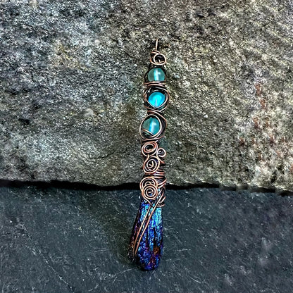 A handmade wire-wrapped pendant featuring three round turquoise-blue beads, copper swirls, and a textured blue and purple kyanite stone at the bottom, displayed against a dark gray stone background.