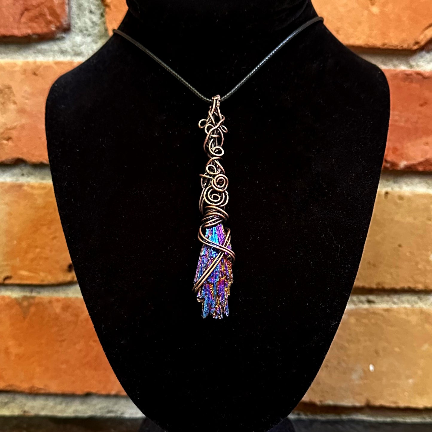 A handmade pendant necklace featuring a copper Celtic knot design wrapping around an iridescent kyanite stone, displayed on a black velvet mannequin bust against a rustic brick wall background.