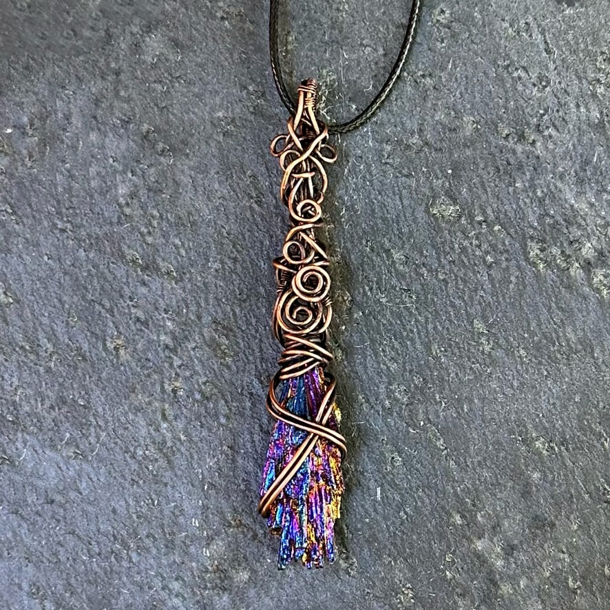 A handmade copper wire pendant necklace featuring an iridescent kyanite stone, resting on a textured gray stone surface.