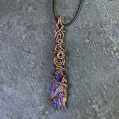 A handmade copper wire pendant necklace featuring an iridescent kyanite stone, resting on a textured gray stone surface.