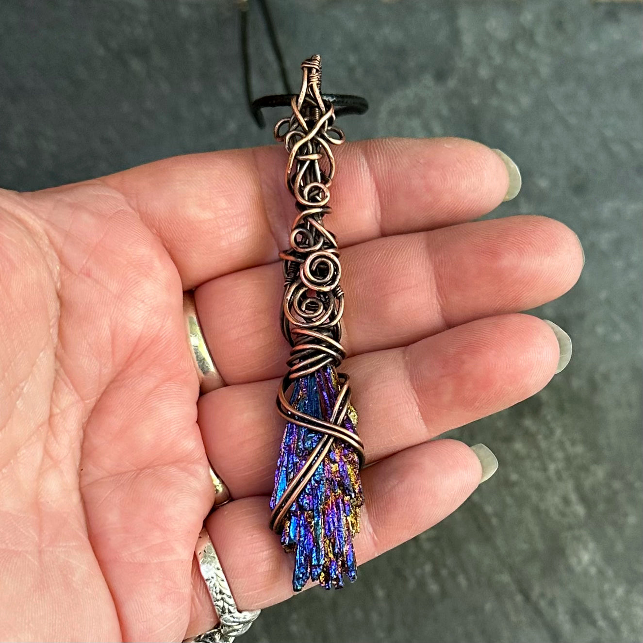 A hand holding a copper-wrapped pendant featuring a blue and purple iridescent kyanite stone, set against a dark textured background.
