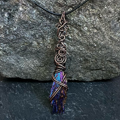 A handmade copper wire kyanite pendant necklace with intricate swirls, hanging against a textured gray stone background.