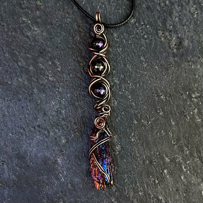 A handmade wire-wrapped pendant necklace featuring a dark iridescent kyanite stone with copper-toned wrapping and dark gemstone beads, displayed on a dark gray slate surface.