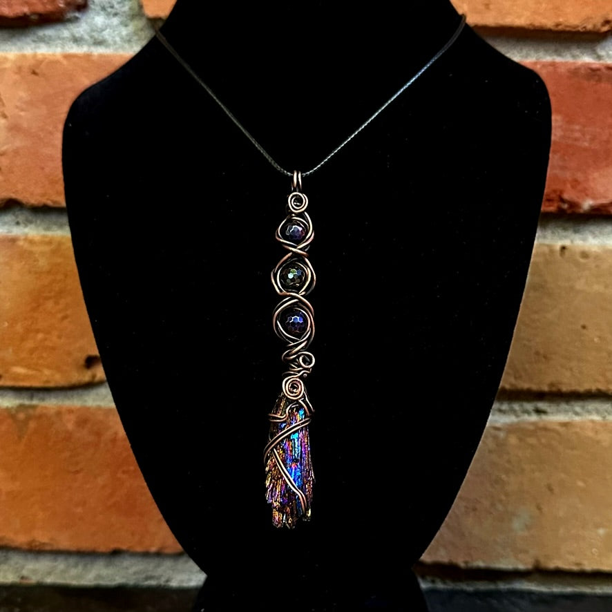 A handmade copper wire-wrapped pendant necklace with iridescent purple beads and a kyanite stone, displayed on a black velvet jewelry bust against a rustic brick wall background.