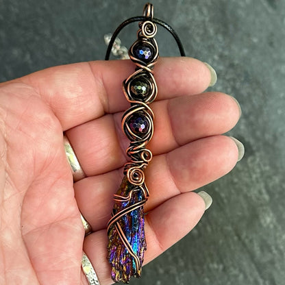 A hand holding a handmade copper wire pendant featuring three iridescent gemstone beads and an iridescent kyanite crystal at the bottom, set against a dark gray textured background.