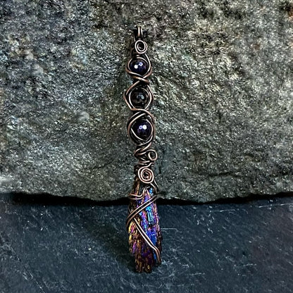 A handmade copper wire-wrapped neckace featuring dark iridescent beads and a purple iridescent kyanite stone on a textured stone and slate background.