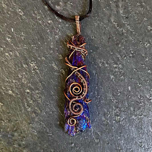 A handmade copper wire-wrapped iridescent purple and blue kyanite stone on a black cord, displayed on a textured gray slate surface.