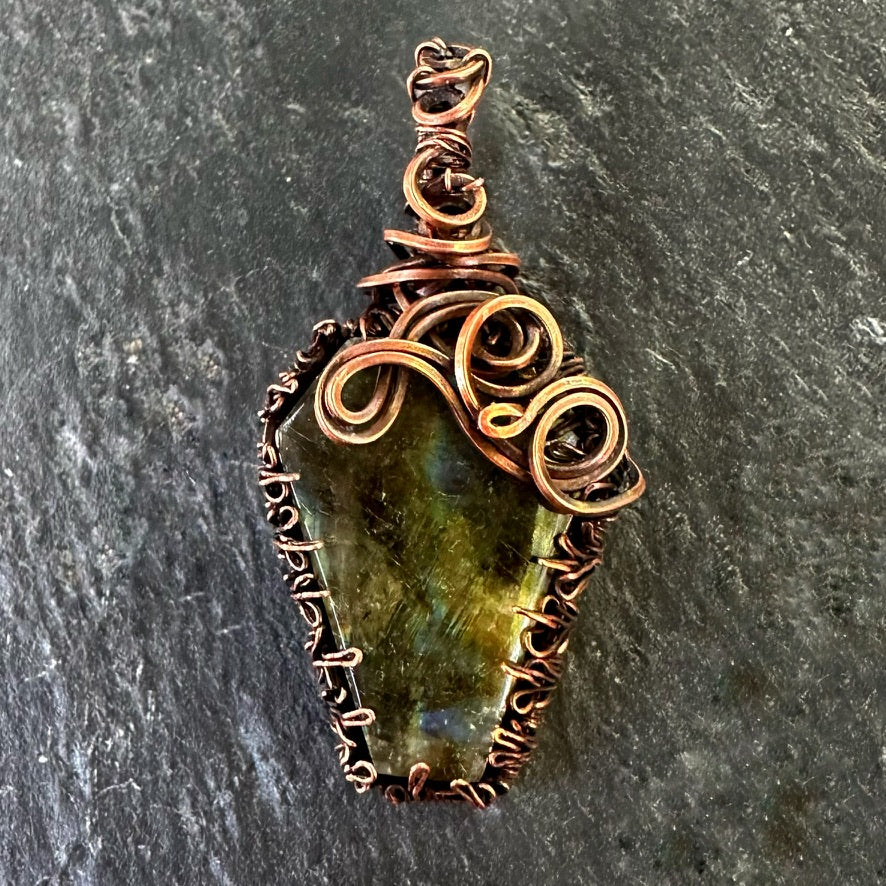 A handmade copper wire-wrapped pendant featuring a coffin shaped labradorite gemstone with iridescent qualities, displayed on a dark textured slate background.