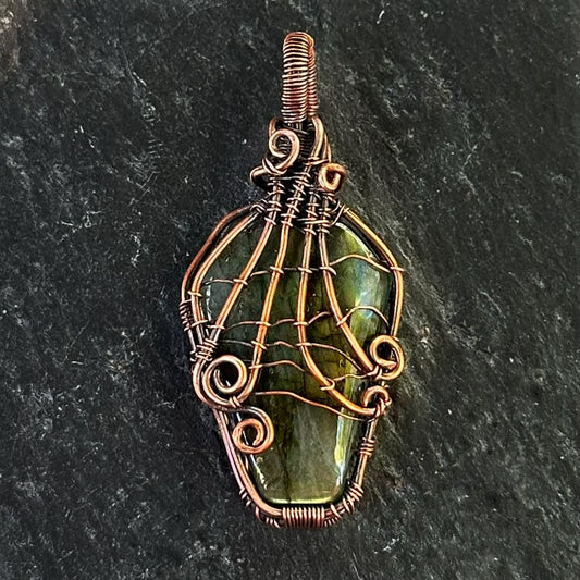 A handmade copper wire-wrapped pendant featuring a coffin shaped labradorite stone with a spiderweb design, displayed on a dark textured slate background.